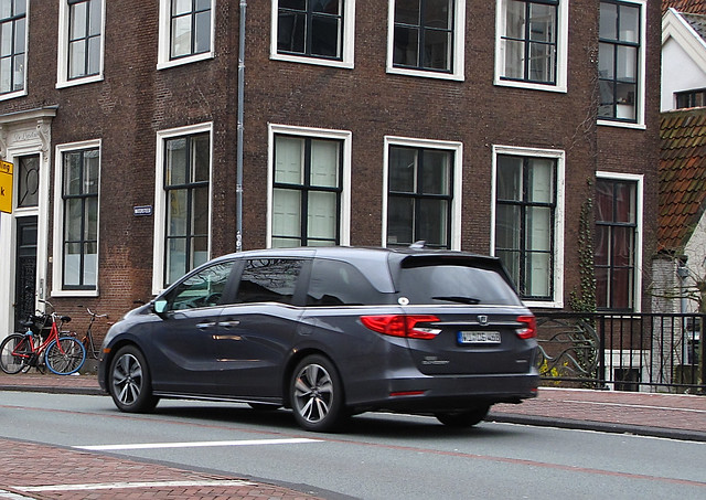 a grey honda odyssey driving through a city in front of a brown building with white windows
